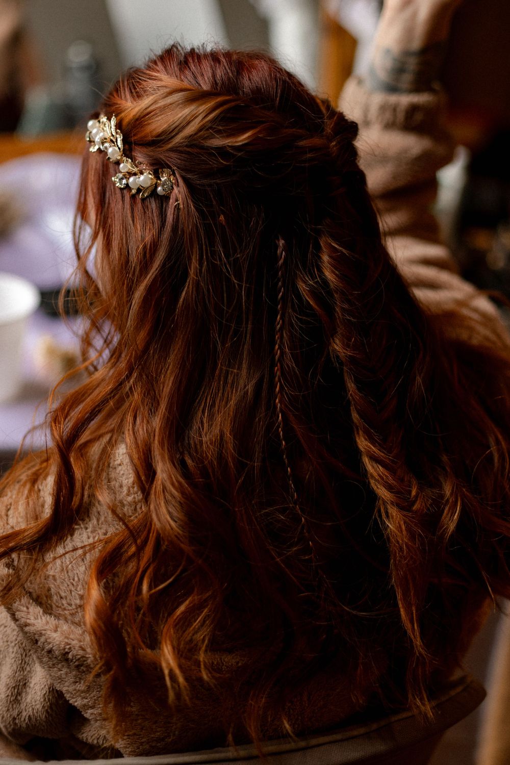 Back view of long auburn wavy hair styled in a half-up look with a delicate pearl and gold floral hair accessory.