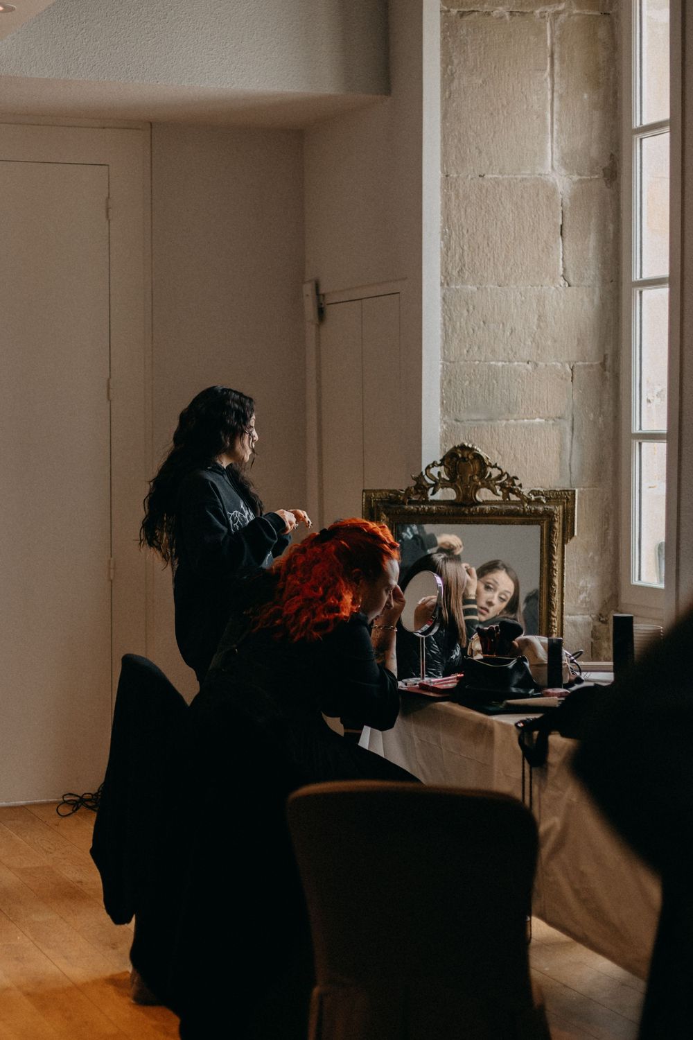 Bride getting ready as a hair stylist works beside an ornate gold mirror in a bright stone room with wooden floors.