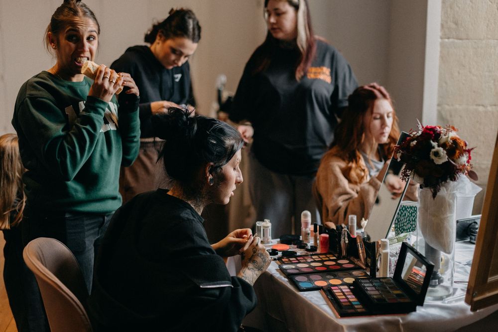 Makeup artists and guests gather around a table of eyeshadow palettes and cosmetics while a woman eats a snack in a bright room.
