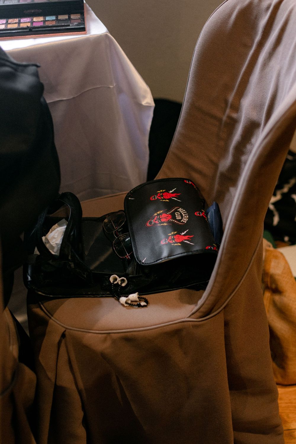 Black handbag with red Berserk-inspired print resting on a taupe covered chair beside a table with makeup palette.