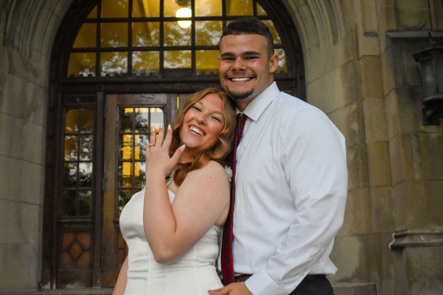 Happy couple poses in front of gothic arch doorway, woman showing engagement ring, man in white shirt and red tie.