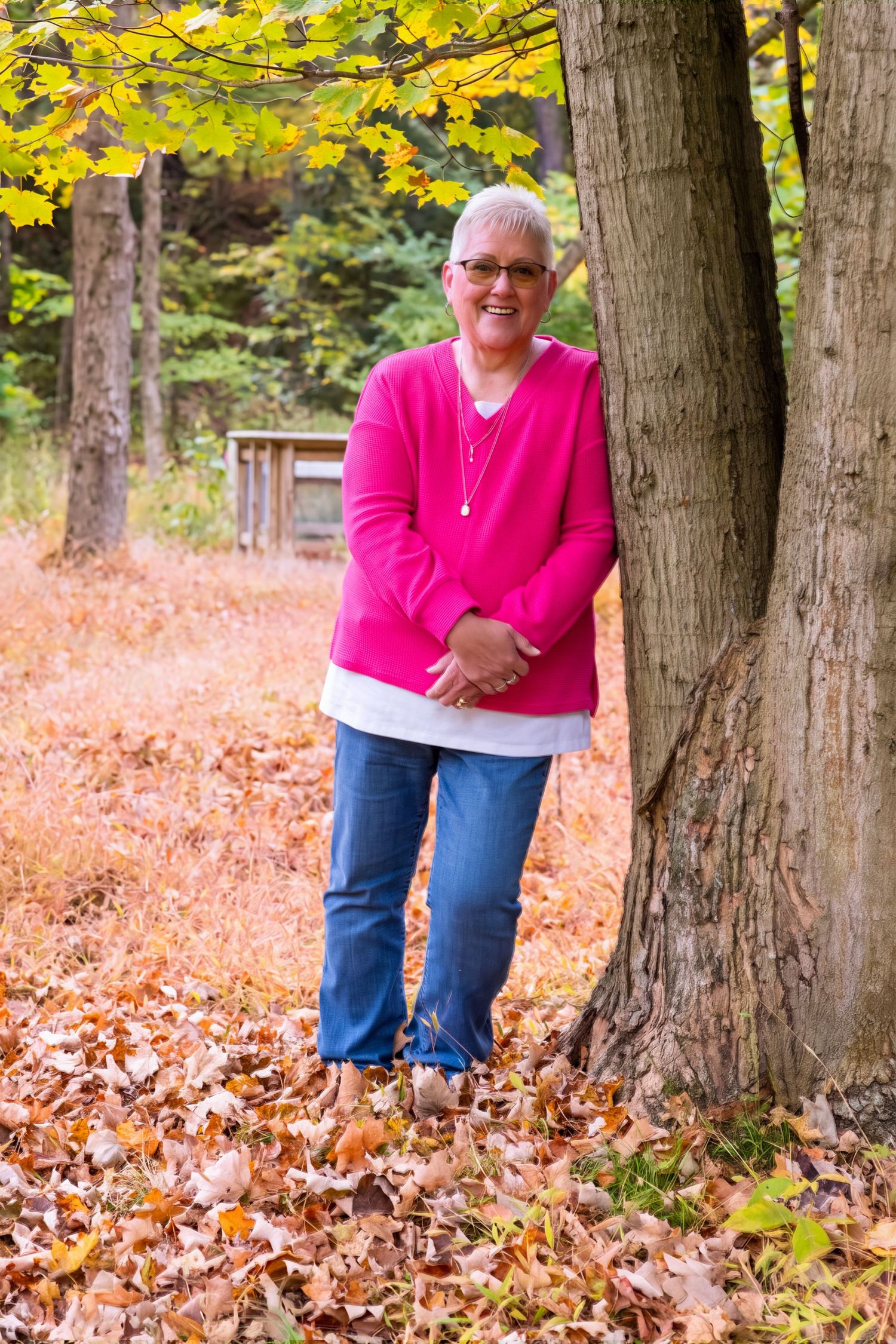 Woman in pink sweater and jeans leaning against a tree surrounded by fall foliage in a forest setting.