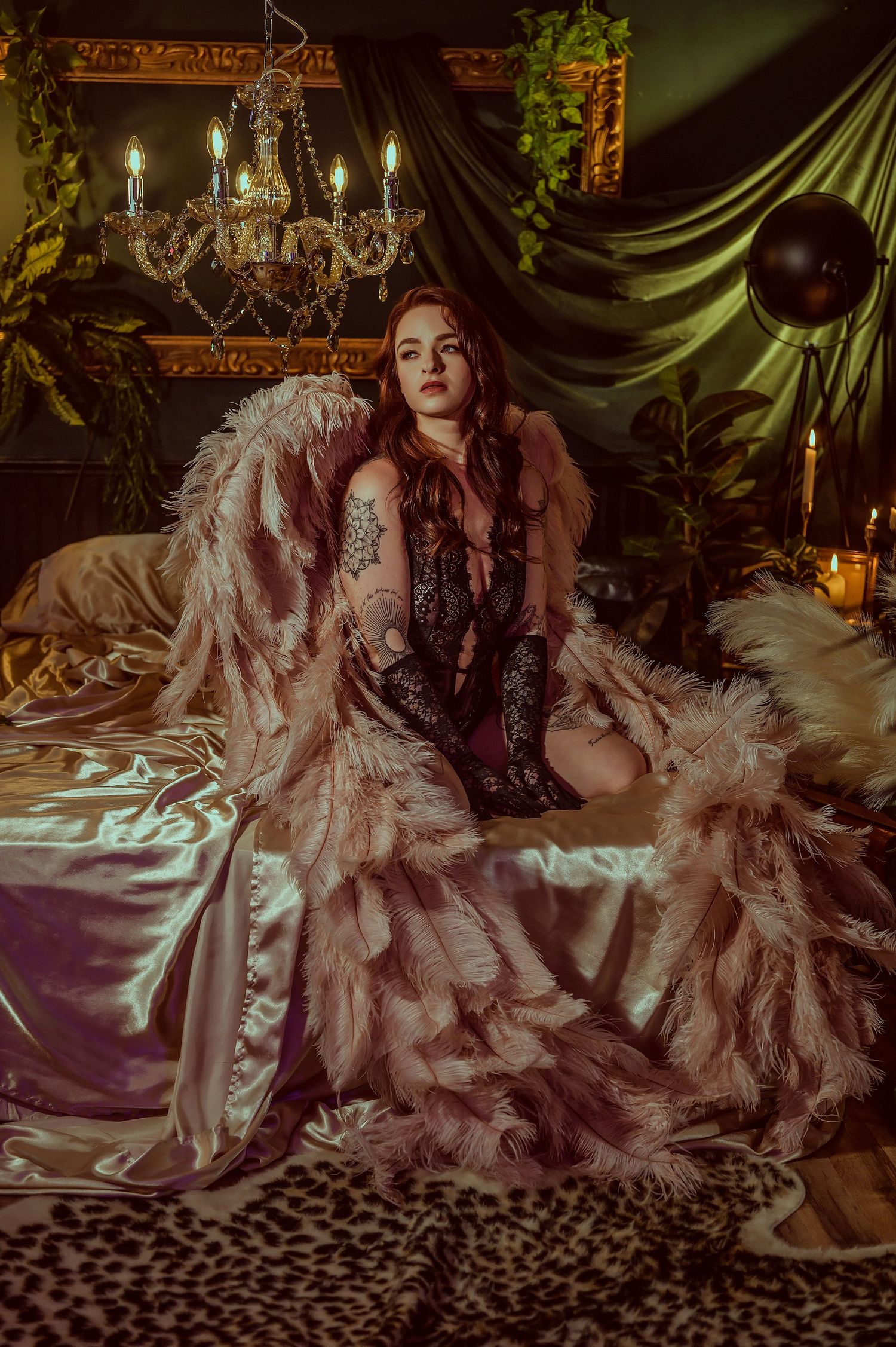 A woman in a pink feathered robe sits on a satin bed in a moody, candlelit boudoir with a chandelier.