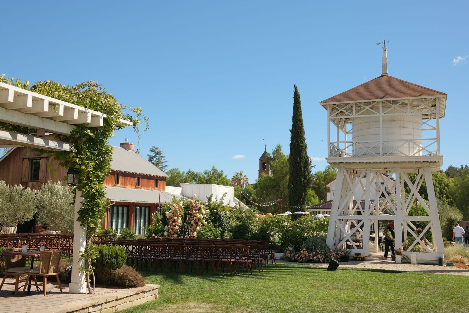 Scenic winery venue with white water tower, manicured lawn, and outdoor seating amid lush gardens on a sunny day.