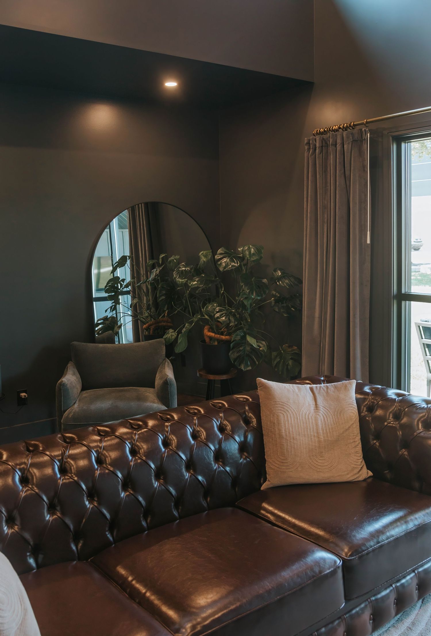 Dark moody living room with tufted brown leather Chesterfield sofa, arched mirror, velvet chair, and lush indoor plants.