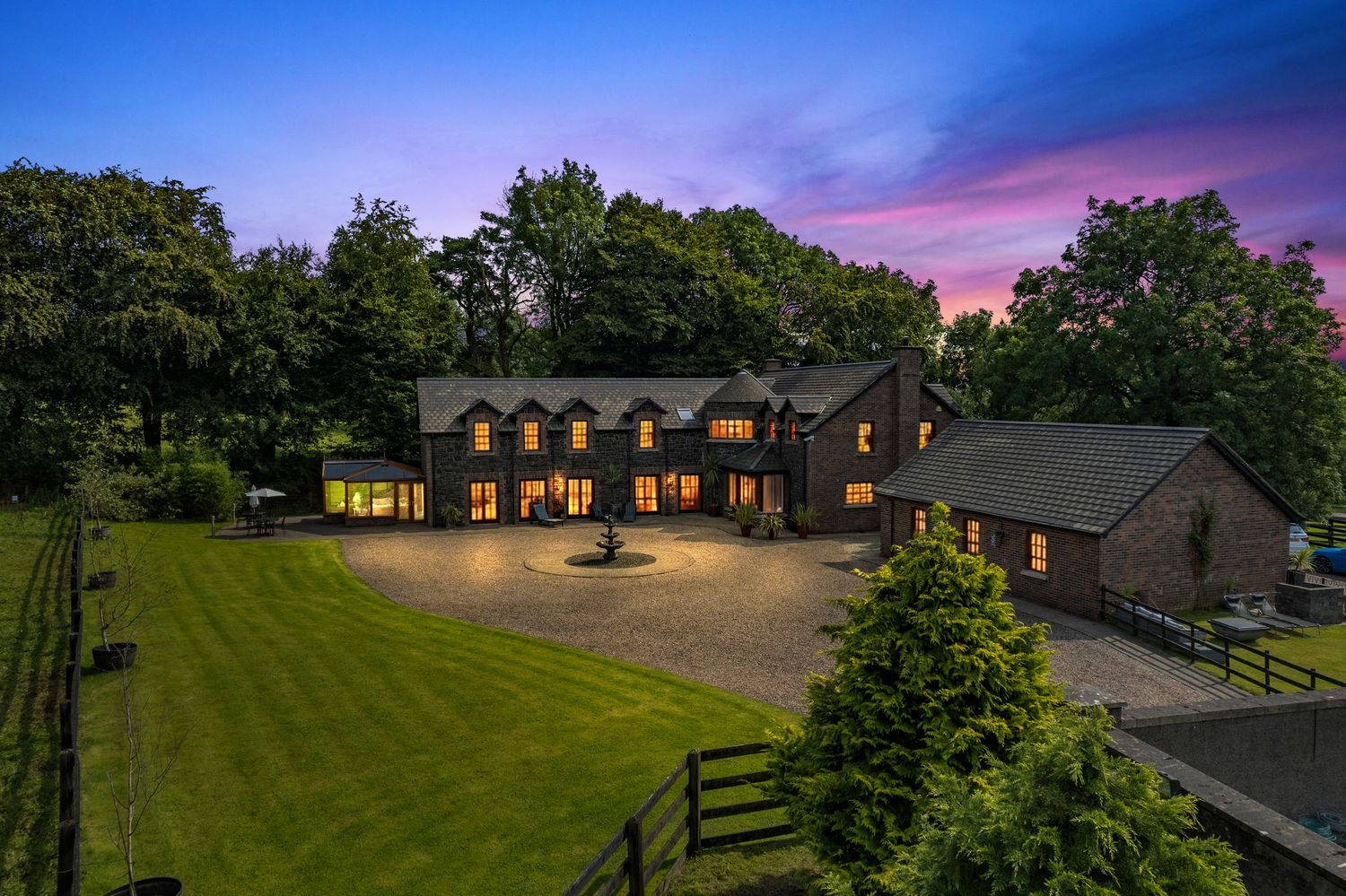 Stunning countryside estate at sunset with manicured lawns, gravel driveway, and warm glowing windows.