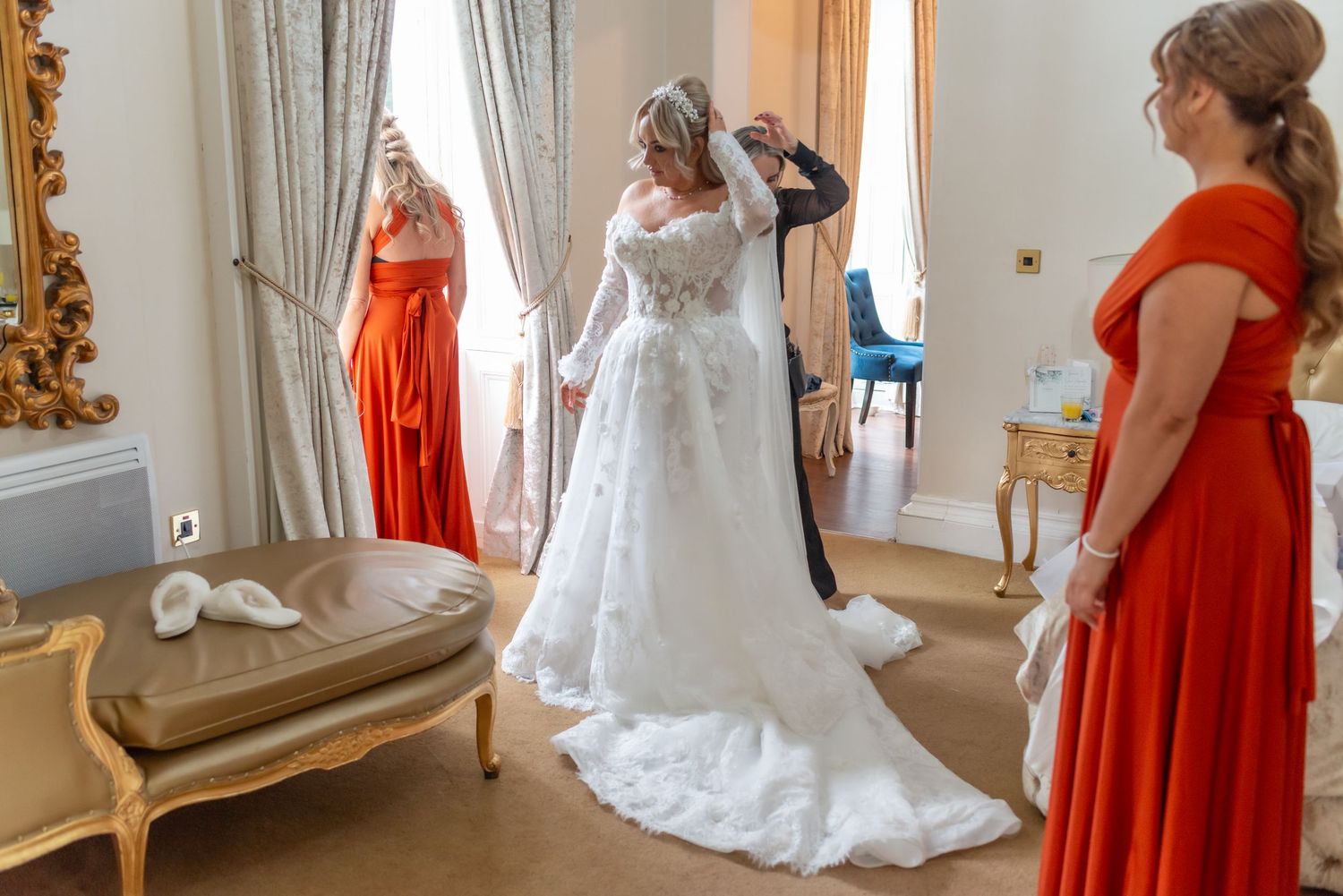 Bride in lace wedding gown getting veil adjusted by helper, with bridesmaids in orange dresses in elegant room at Rainhill Hall Hotel