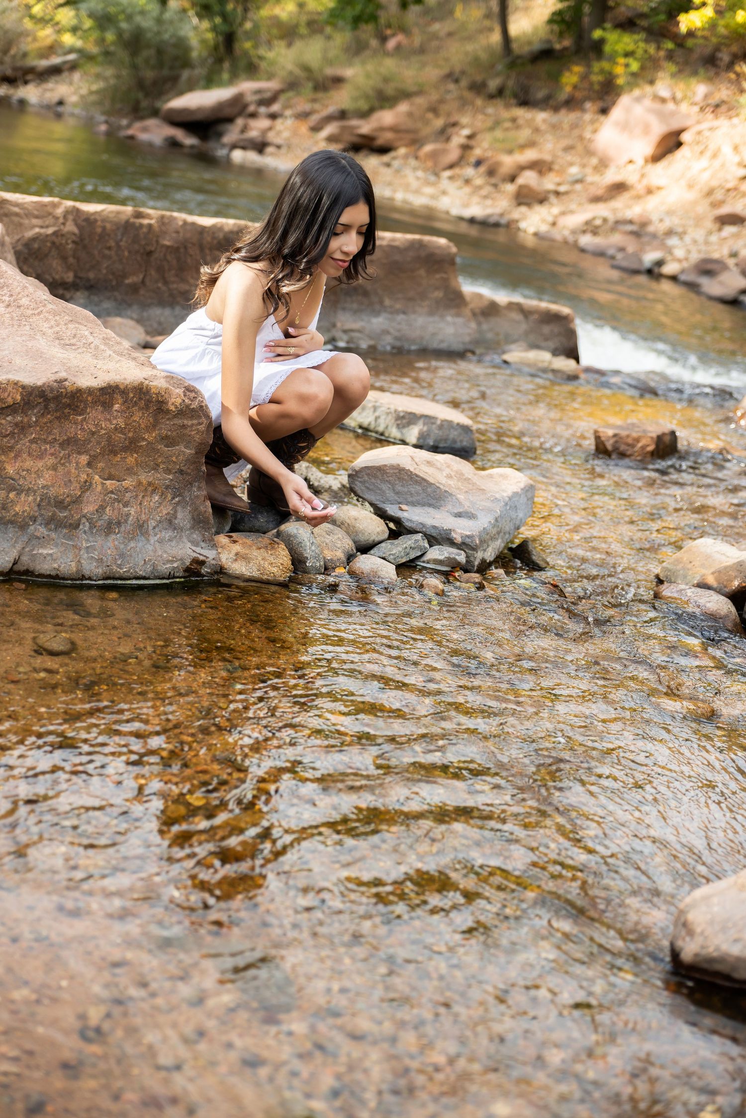 Young woman in white sundress and cowboy boots standing on rocky path by a serene creek surrounded by lush green trees.