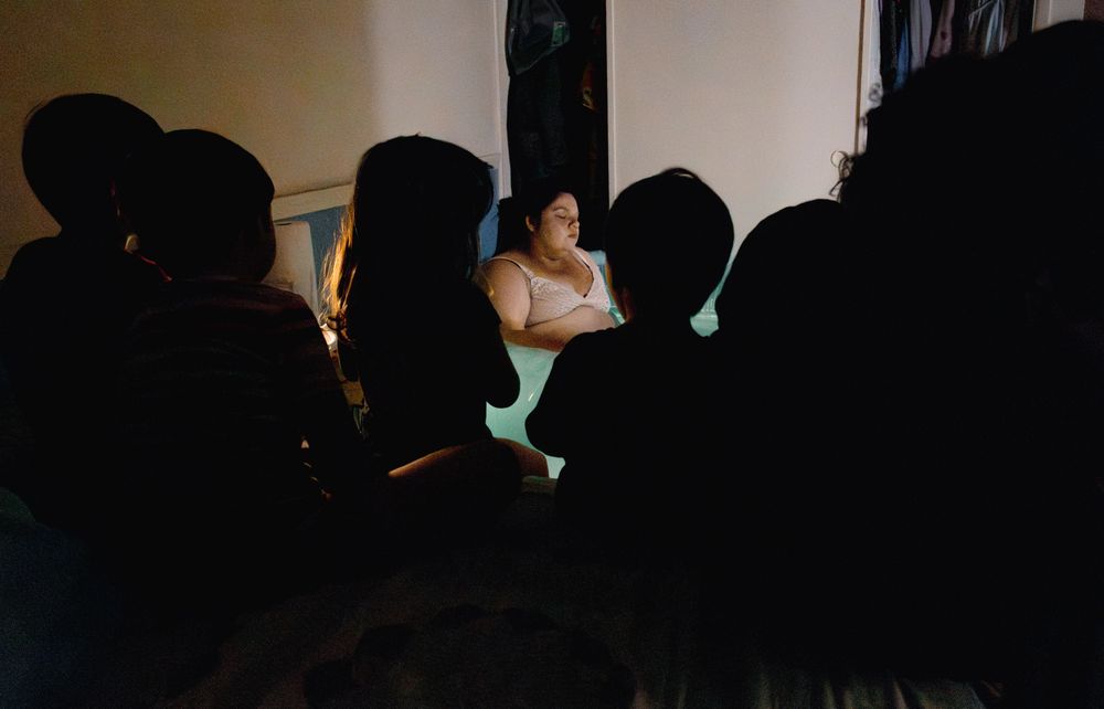 Woman sitting in a dimly lit room surrounded by silhouetted audience members during an intimate gathering.