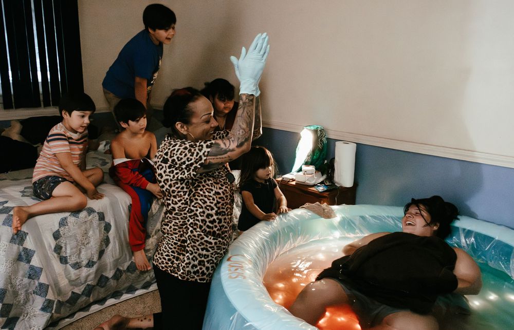 A midwife in gloves assists a woman in an inflatable birth pool during a home water birth with family present.