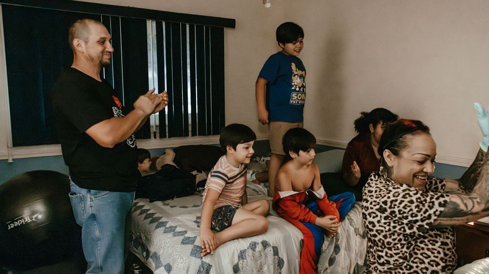 Family gathering in a bedroom with adults and children sitting on a bed, laughing and enjoying time together indoors.