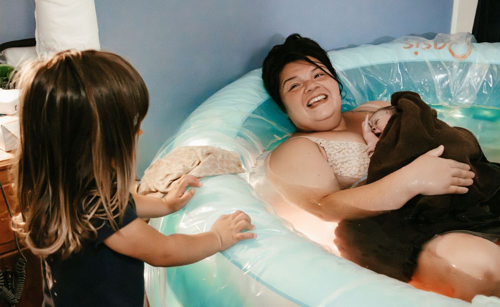 Woman laboring in birth pool smiles while midwife assists during water birth.