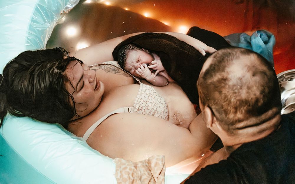 Mother holds newborn moments after water birth as partner looks on lovingly.
