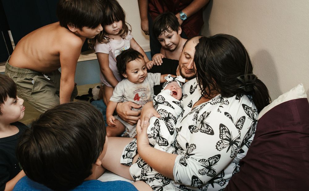 A woman in a butterfly-print top holds a newborn while surrounded by several young children in a warmly lit room.