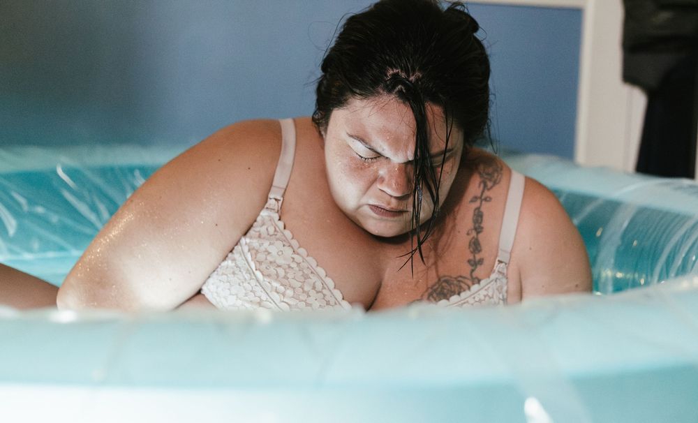 Woman in lace bra labors in inflatable birth pool during home water birth.