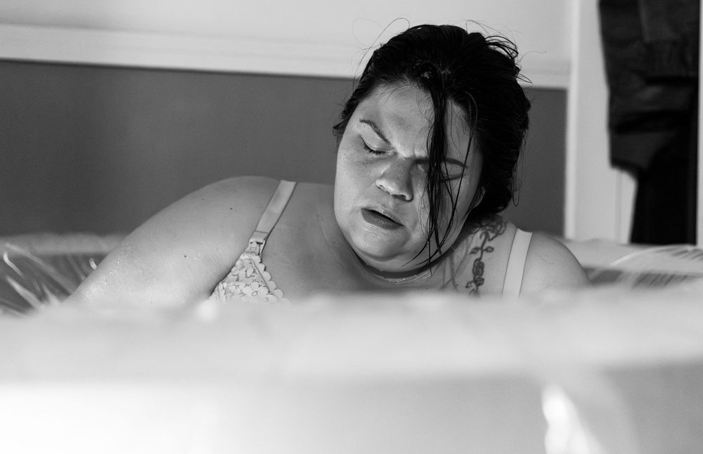 Black and white photo of a woman in a birthing pool during labor, eyes closed, appearing to be in active childbirth.