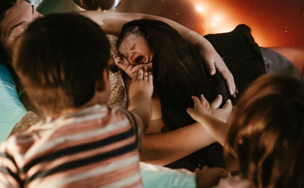 Emotional birth moment as a woman holds her newborn surrounded by loved ones in warm ambient light.