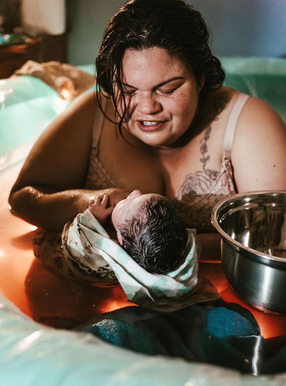 A new mother smiles with joy while holding her newborn baby wrapped in a towel during a water birth.