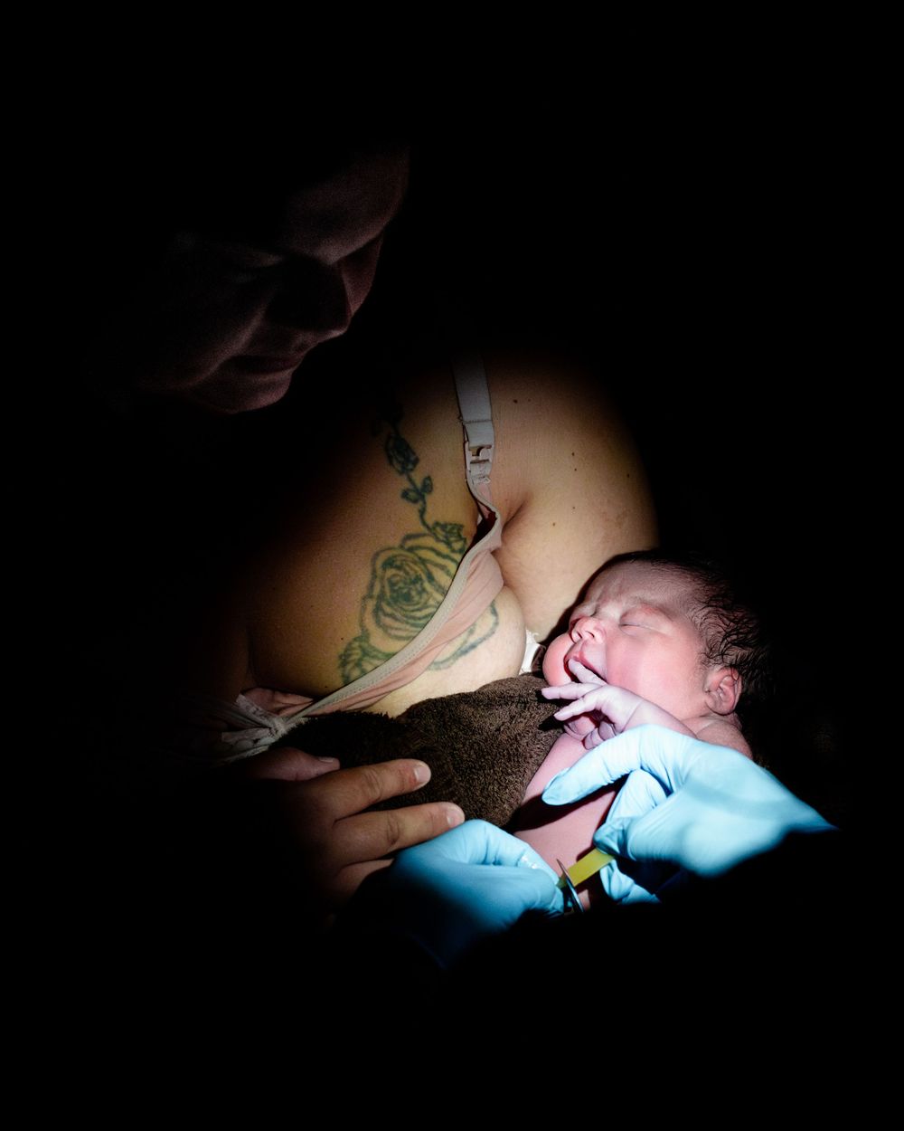 A mother holds her newborn baby moments after birth in a dimly lit room, with a medical professional in blue gloves assisting.