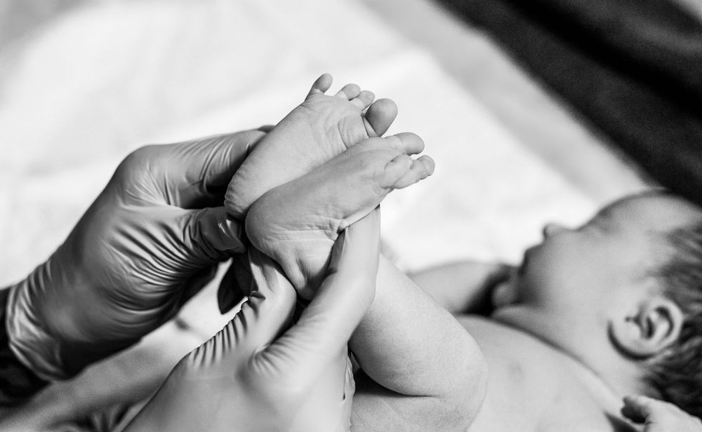 Black and white photo of a newborn baby's feet being held by gloved medical hands during a checkup.