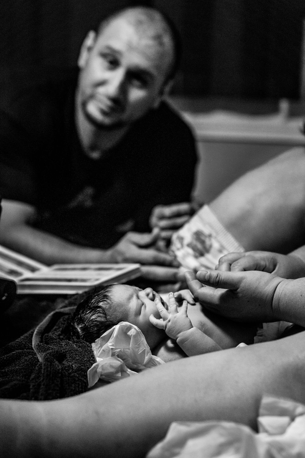 Black and white photo of a newborn baby being held by hands while a smiling man looks on in a hospital room.