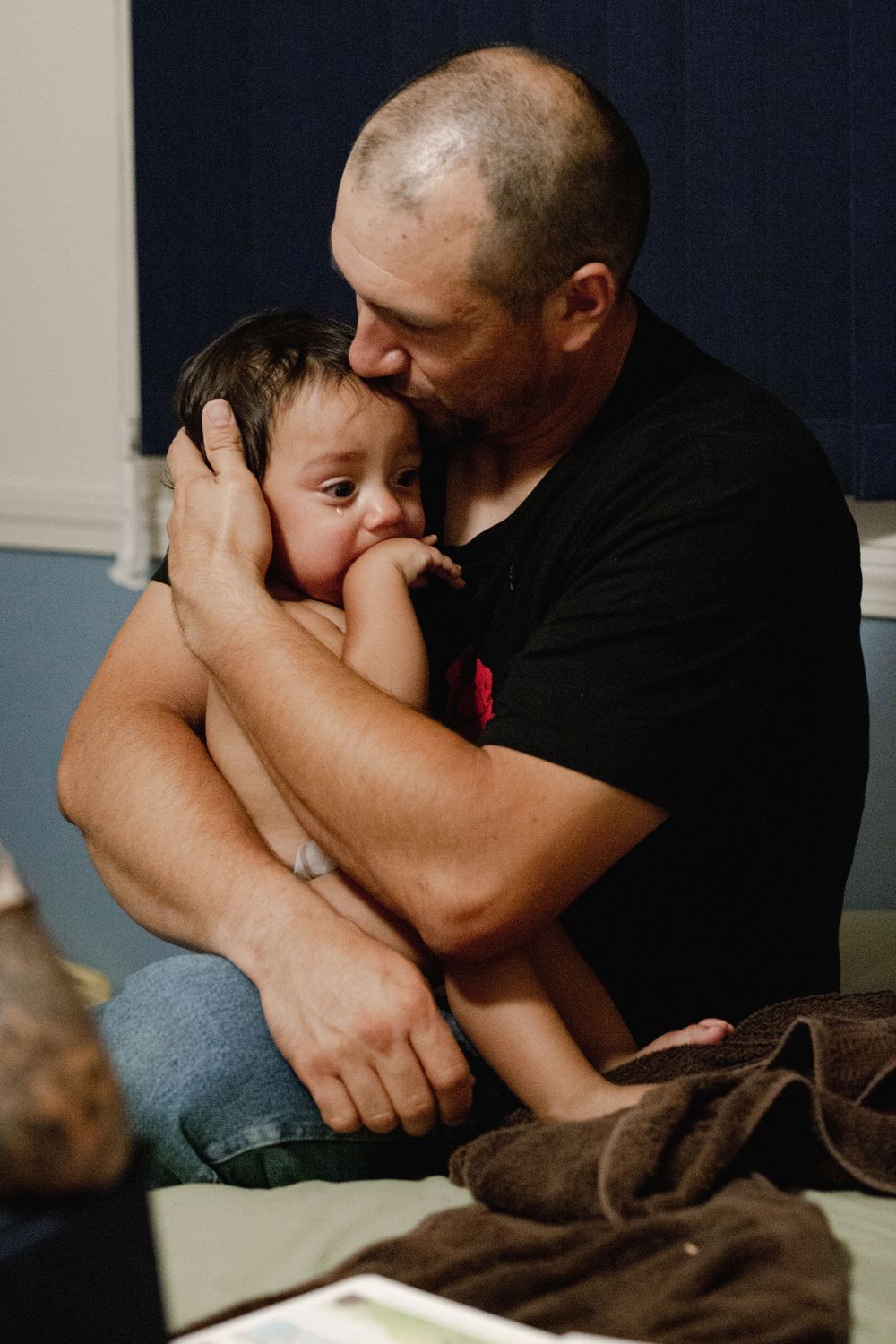A bald man in a black shirt tenderly kisses and hugs a young toddler sitting on his lap.