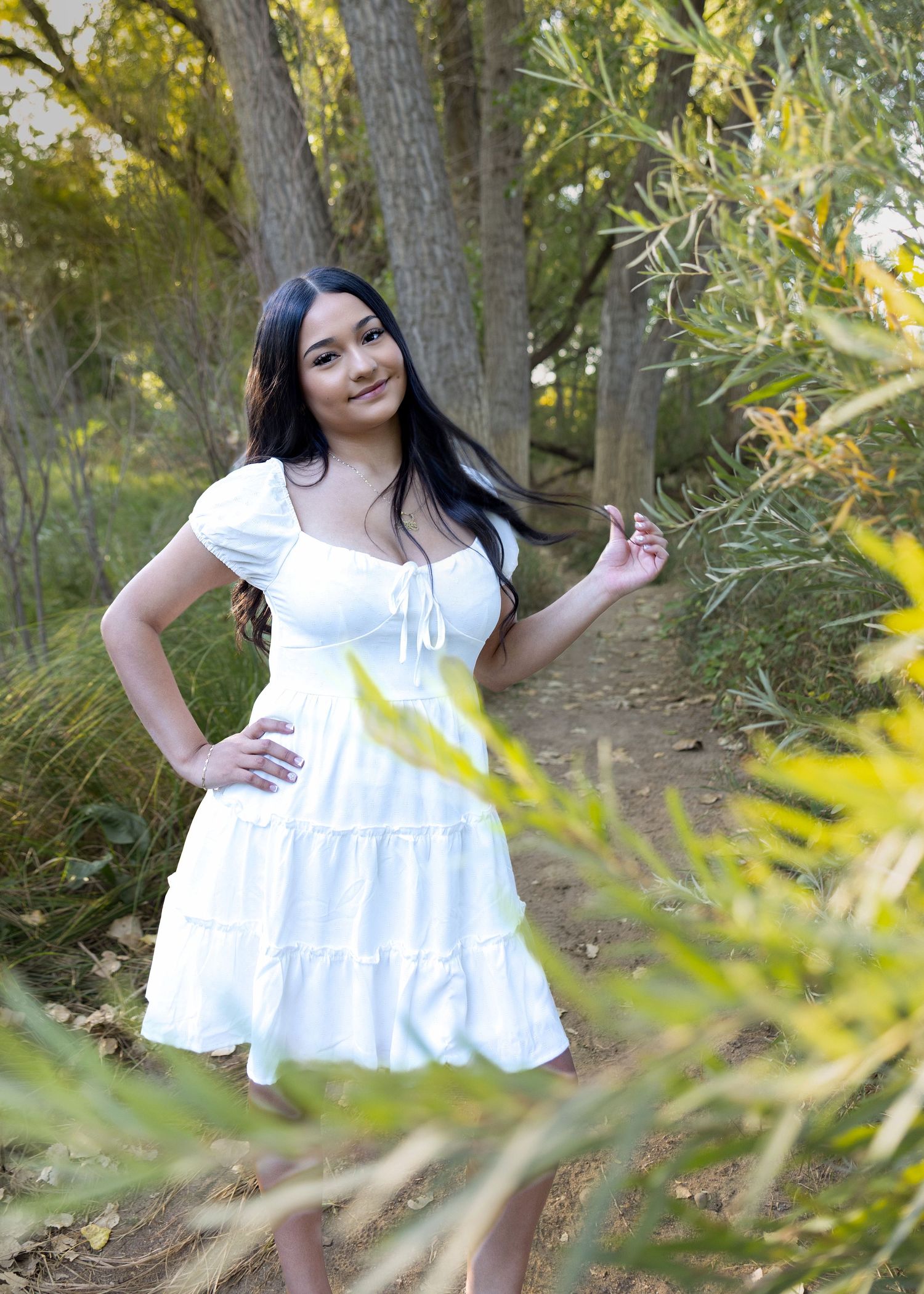 Young woman in white dress poses gracefully among lush green trees on a scenic outdoor path.