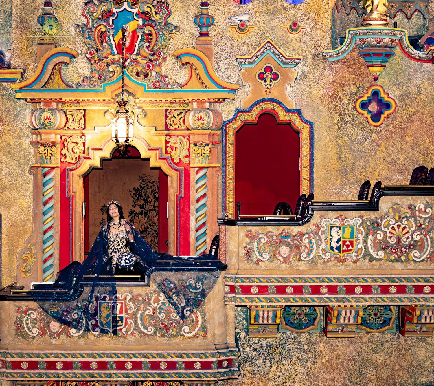 Woman in blue gown poses in ornate, colorful theater interior with gilded arches and decorative murals.