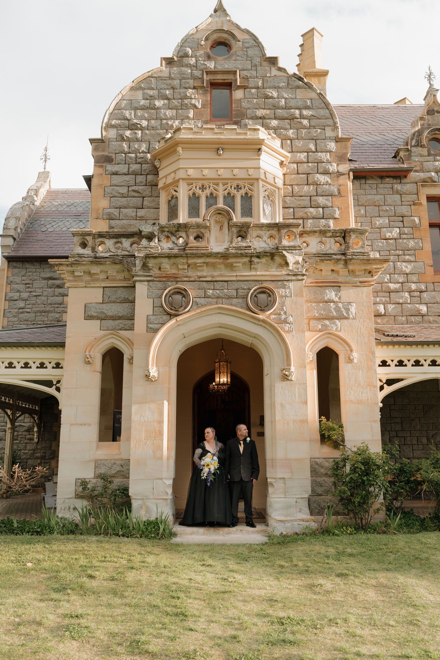 Couple in black wedding attire standing in ornate stone castle entrance with arched doorway and Gothic architecture.