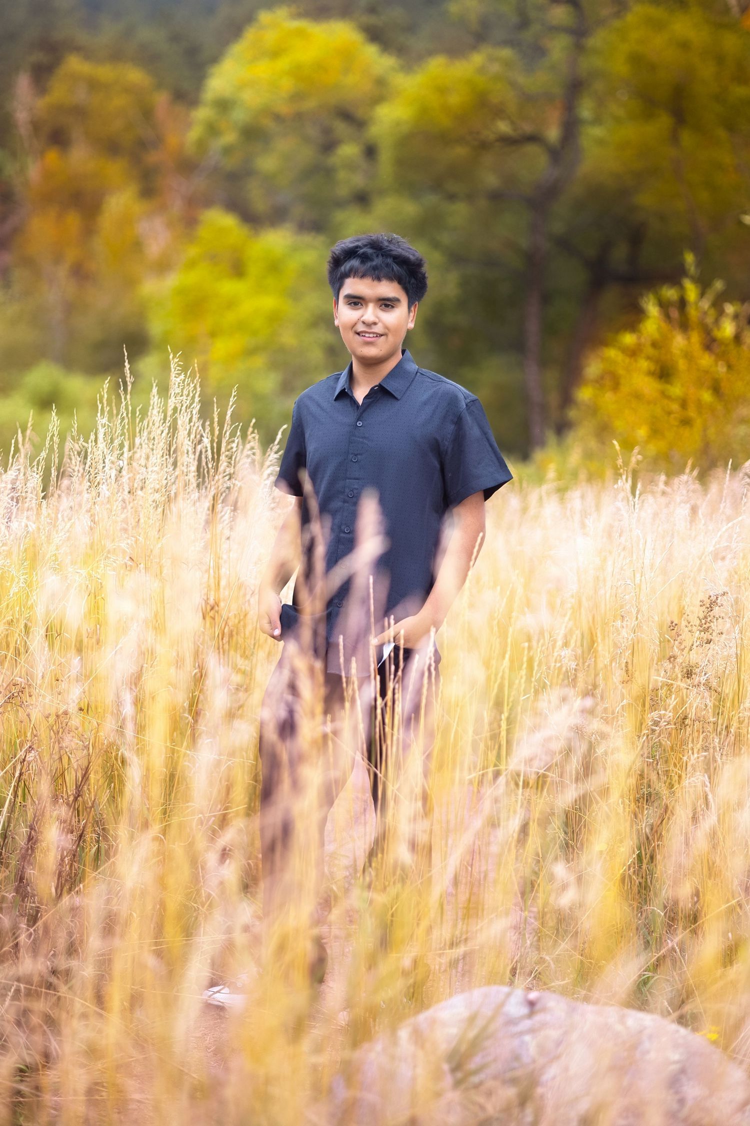 Young man in dark shirt standing in golden tall grass field with colorful autumn trees in background.
