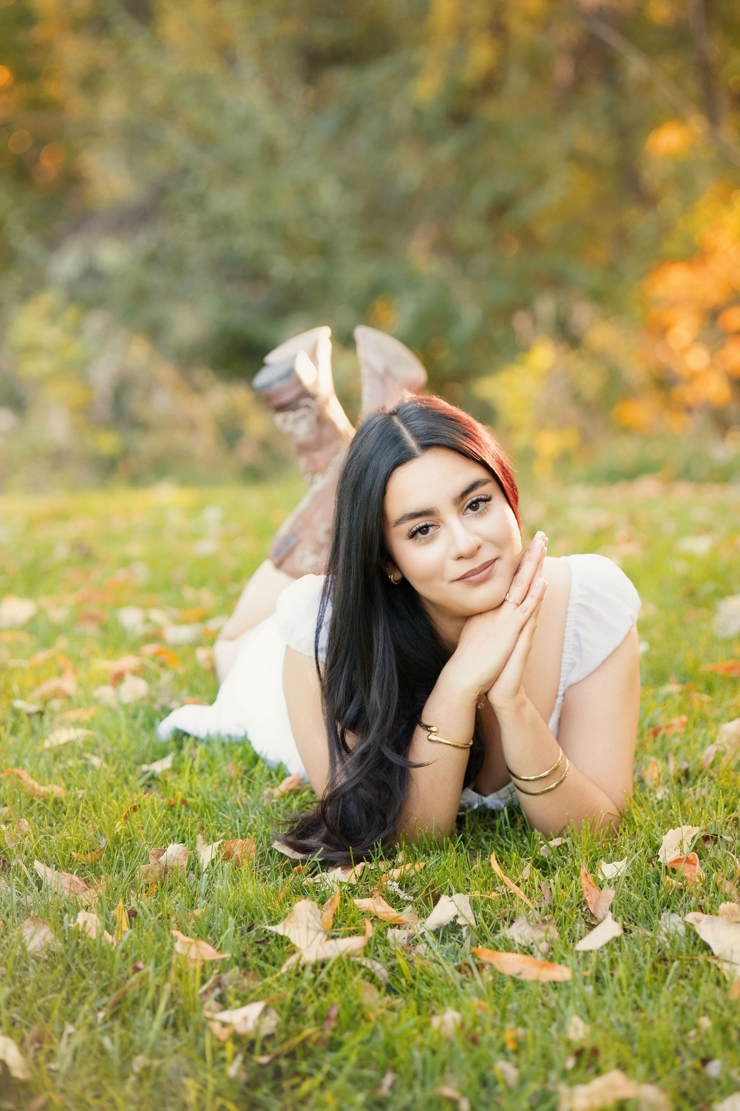 Young Asian woman with long dark hair lying on green grass in autumn park, smiling softly with hands under chin.