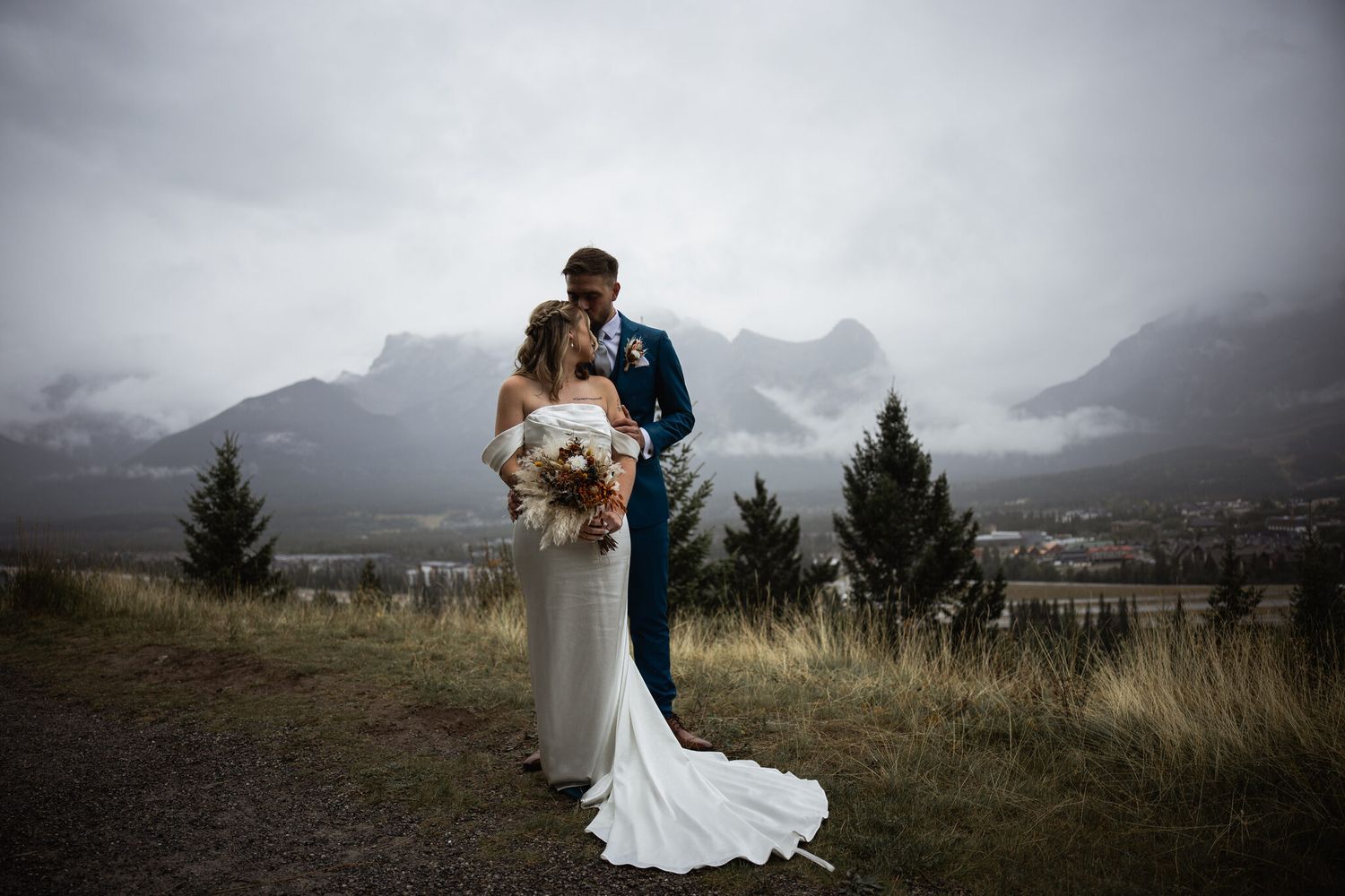 Bride and groom embrace on a misty mountain overlook, she in a white gown holding a bouquet, he in a navy suit.