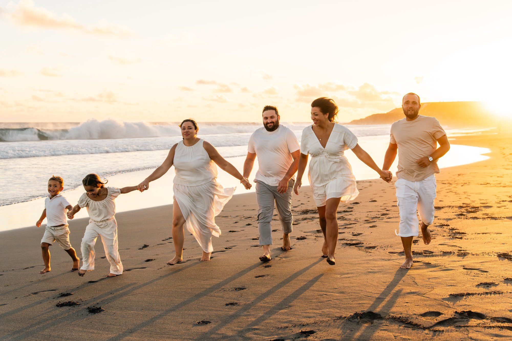 Happy family running on beach at sunset in white outfits, waves crashing behind them on a golden shore.