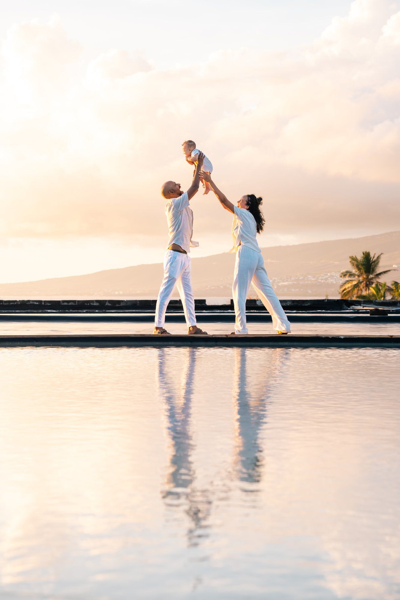 Family in white outfits lifting baby by infinity pool at sunset with ocean and mountains backdrop.