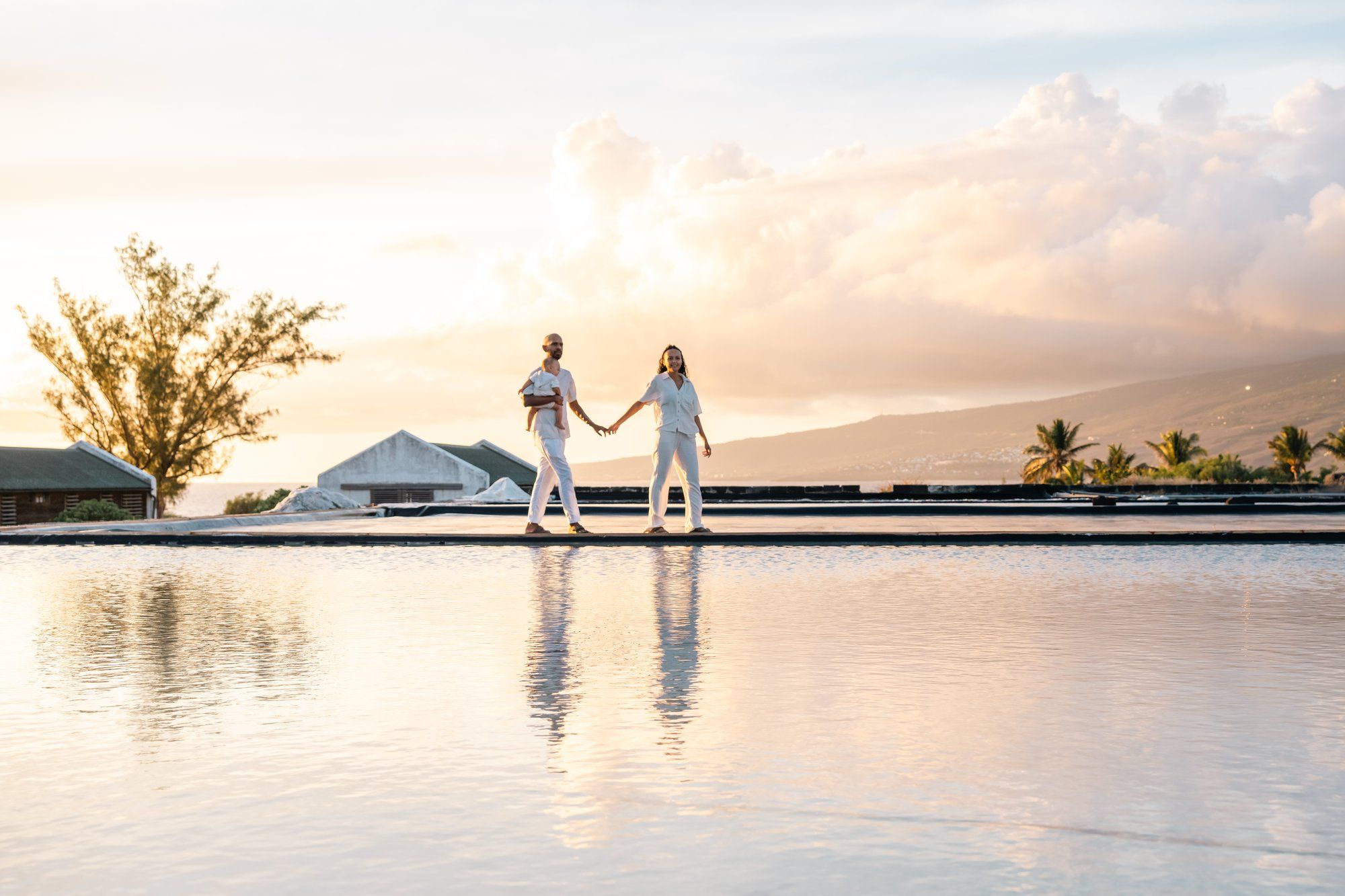 Couple in white holding hands by infinity pool at sunset with tropical landscape and dramatic clouds.