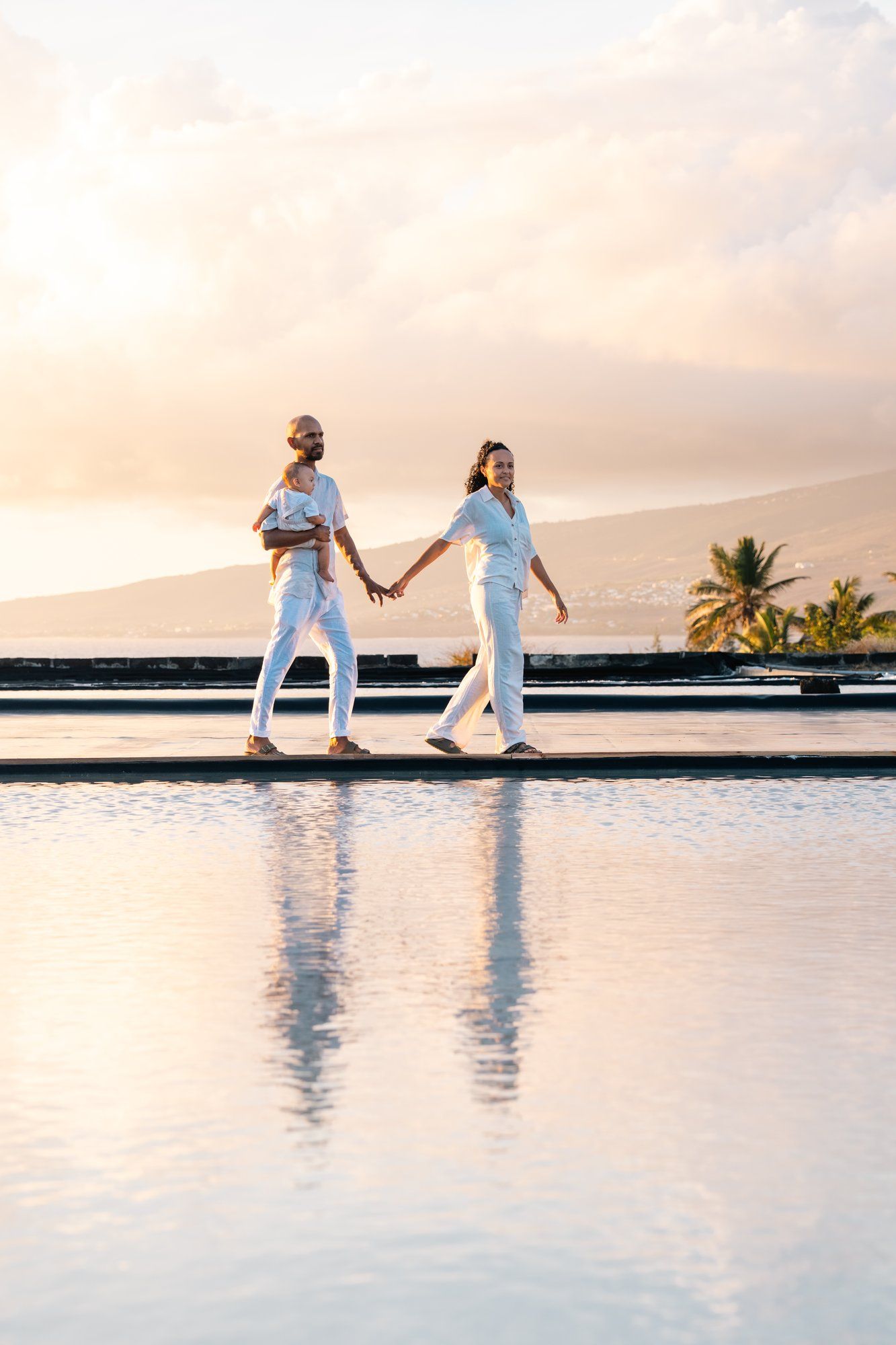 Couple in white outfits holding hands walking by infinity pool at sunset with palm trees and mountains.