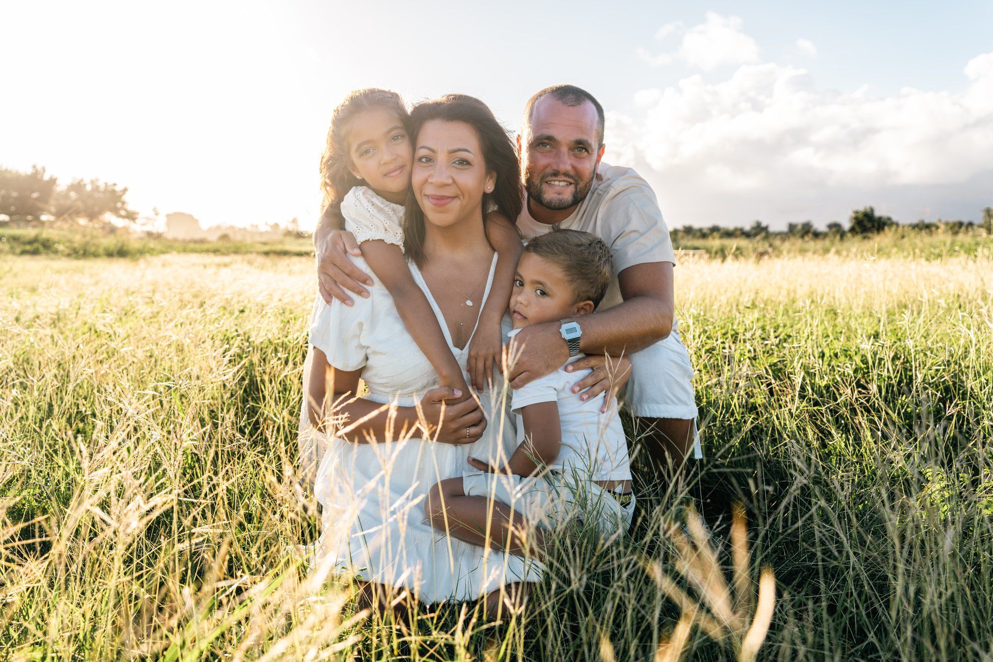 Happy family of four posing together in a sunny golden grass field, parents embracing two young children outdoors.