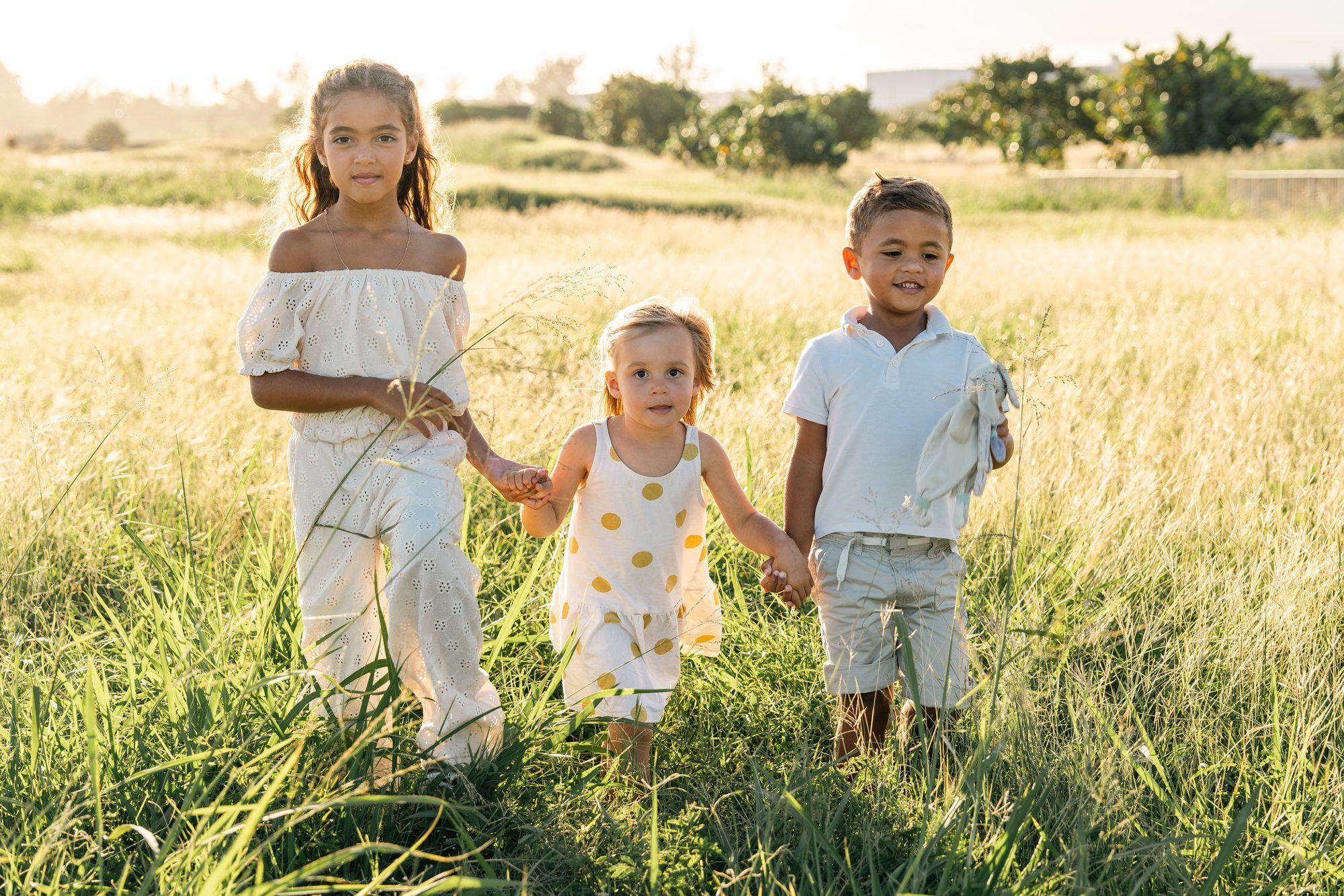 Three children holding hands in a sunny field, two older kids flanking a toddler in yellow dress, golden hour lighting.