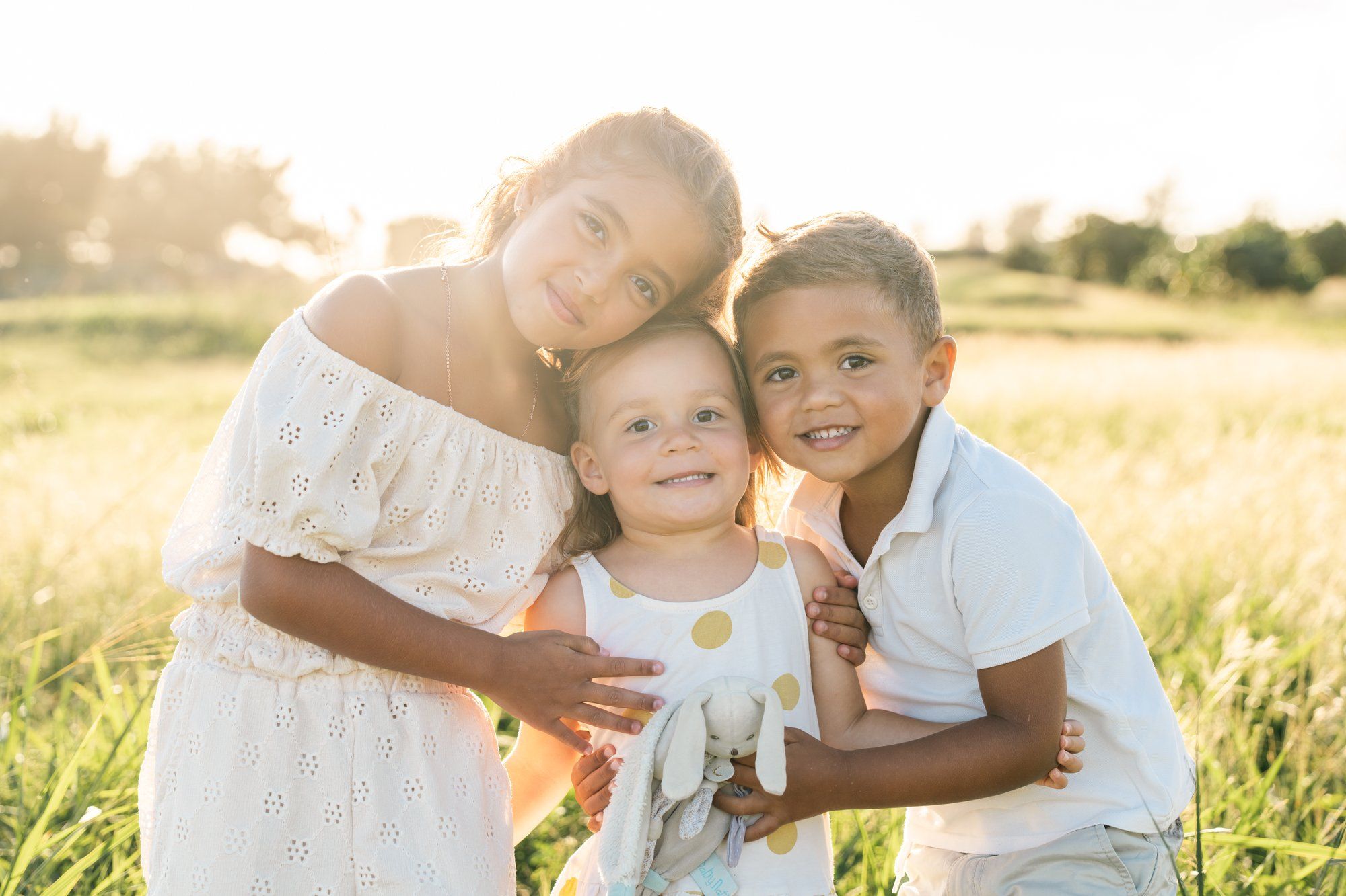 Three happy children posing together in a sunny field, wearing white outfits in a warm golden light.