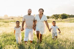 Family of four walking hand-in-hand through a sunny green field, wearing white outfits in a warm golden light.