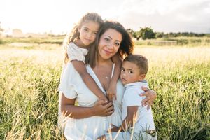 A mother hugs her two young children in a sunlit grassy field, all wearing white in a warm family portrait.