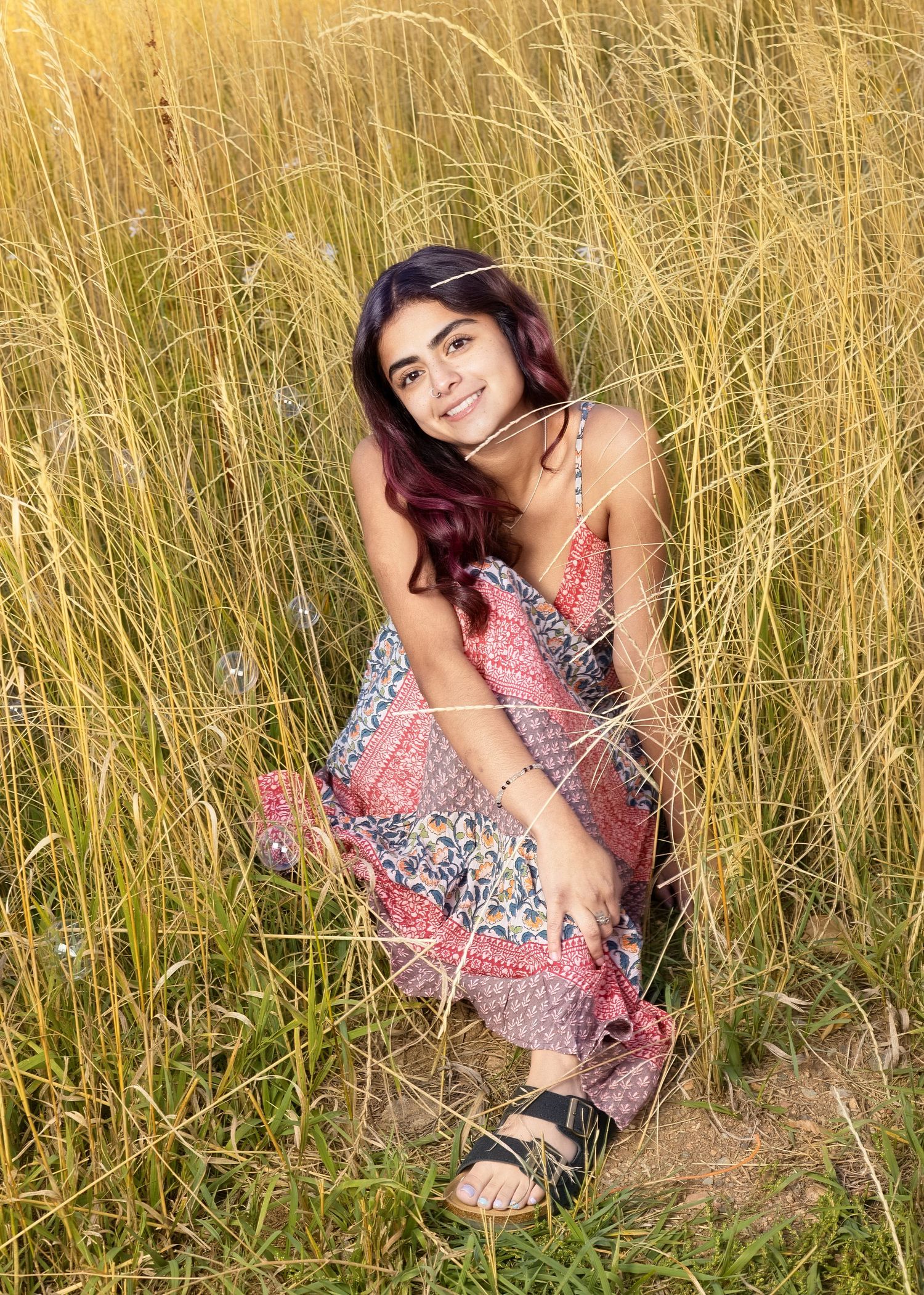 Young woman in floral dress crouching in tall golden grass field, smiling warmly in natural sunlight.