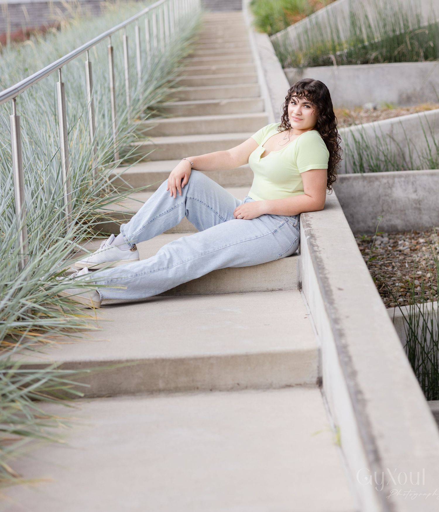 A young woman in a light green crop top and light wash jeans sits relaxed on outdoor steps near ornamental grasses.