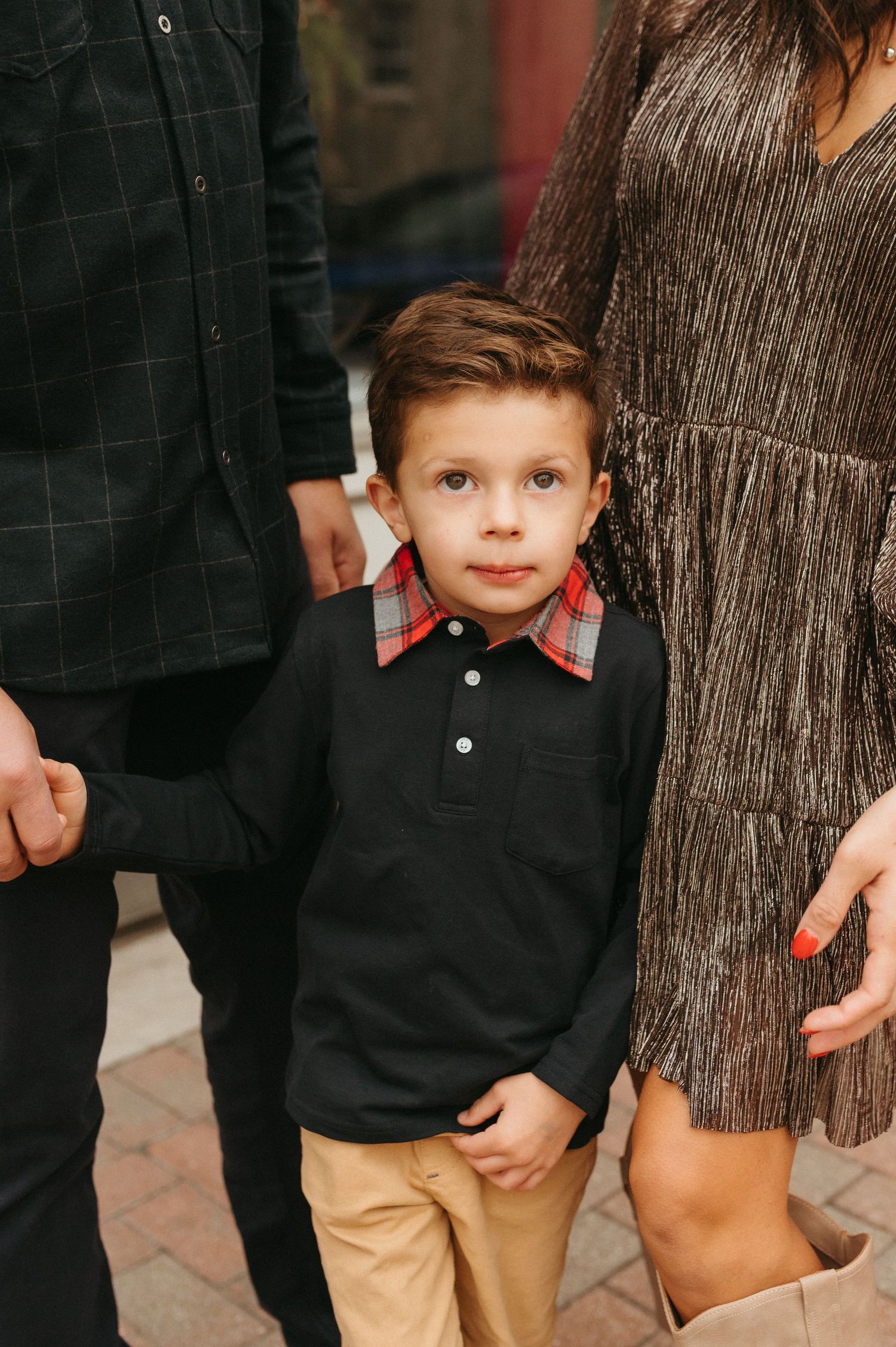A young boy in a black polo shirt with plaid collar and khaki pants stands between two adults during a family photo session.