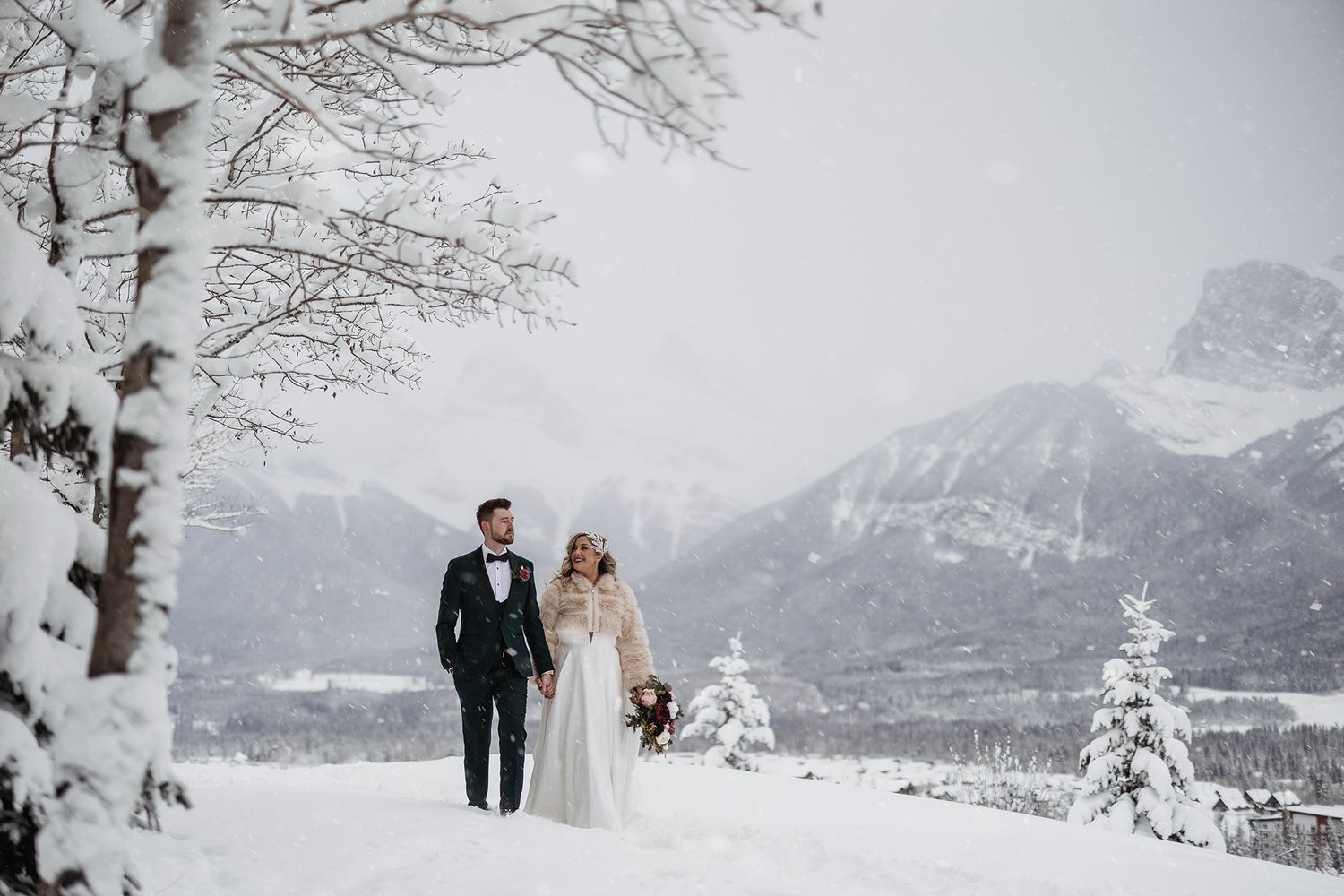Bride and groom walking through snowy winter landscape with snow-covered trees and mountains in background.