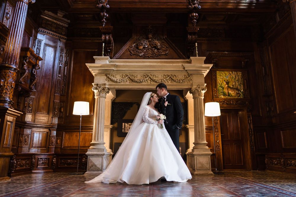 Elegant bride and groom sharing a romantic moment in a grand wood-paneled hall with ornate fireplace.