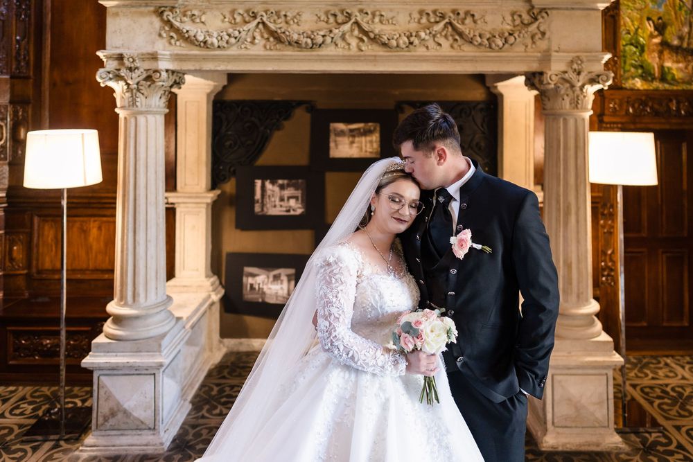 Elegant bride and groom posing together in ornate venue with grand fireplace, bride holding bouquet in lace wedding gown.