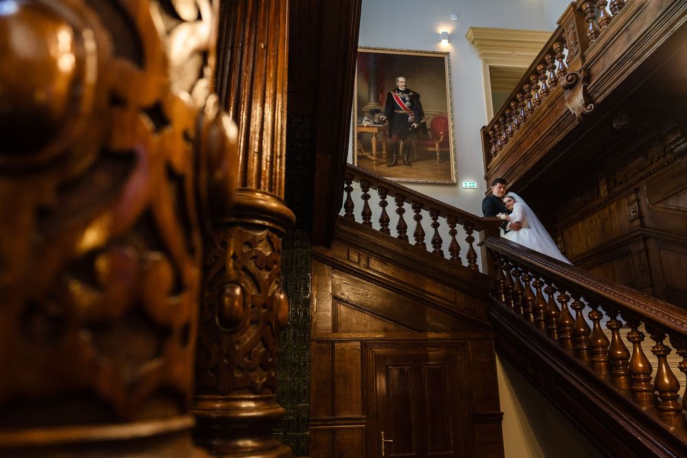 Bride and groom share a romantic moment on an ornate wooden staircase beside a classical portrait painting.
