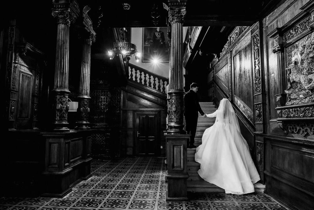 Black and white wedding photo of bride and groom ascending grand staircase in ornate historic mansion.