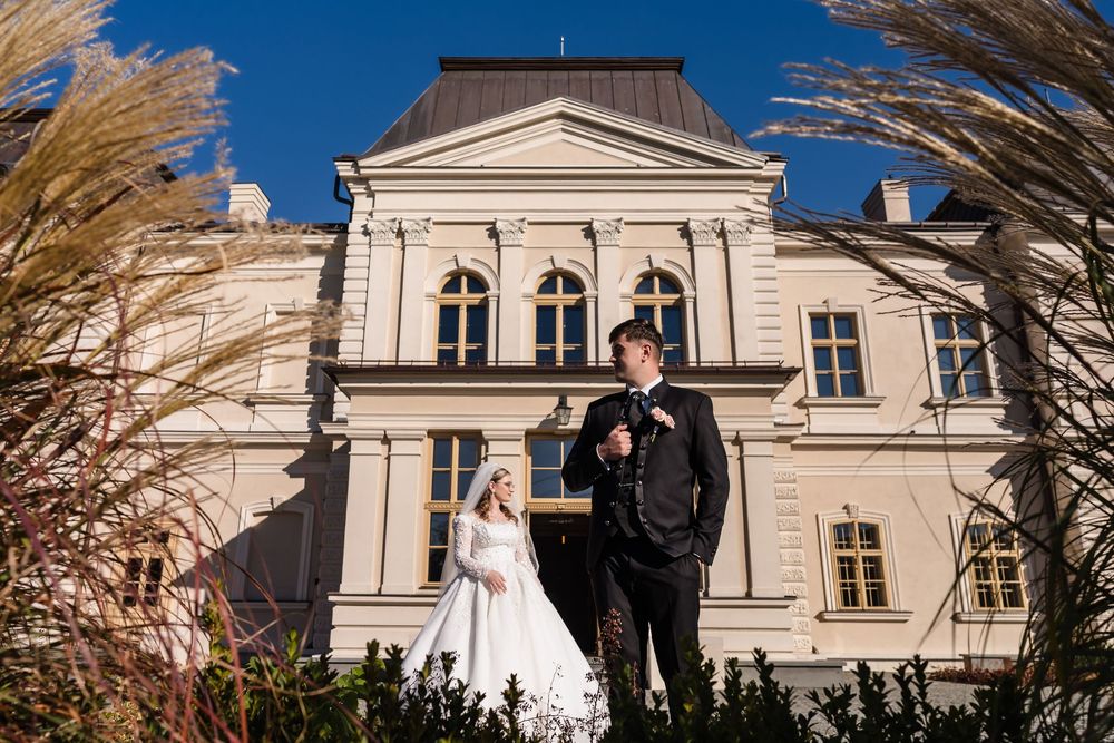 Bride and groom posing in front of a grand historic mansion surrounded by ornamental grasses on a sunny day.